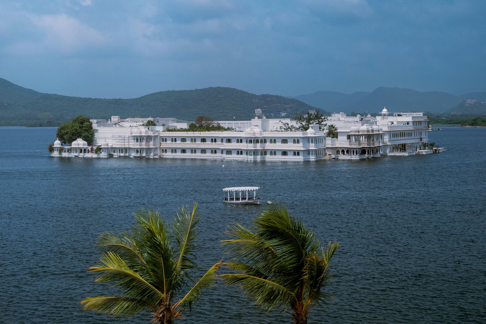 White marble Lake Palace floating on Lake Pichola in Udaipur with the Aravalli hills behind and palm trees in the foreground